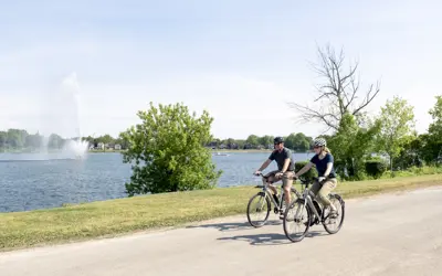 A couple cycling on a roadway; Peterborough's Little Lake is in the background with the Centennial Fountain running - sky is blue, early